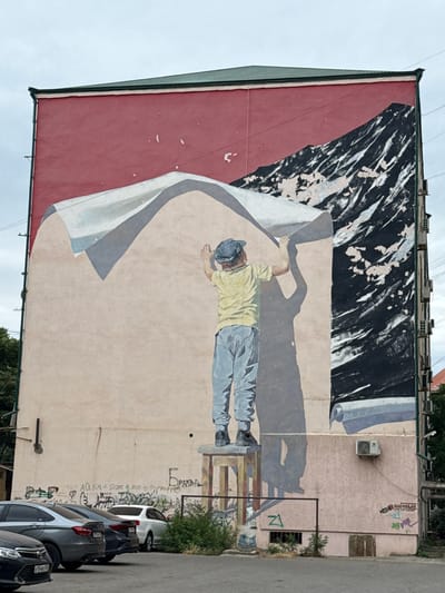 This is a photograph of a mural painted on the side of a building. The mural depicts a man in a yellow shirt and blue pants, wearing a blue cap. He is lying down and appears to be unrolling a large sheet of paper or canvas, which is colored a light beige. The top portion of the mural features abstract black and white shapes, possibly representing a torn or peeling surface. The man is casting a shadow on the beige surface.
The scene is set outdoors, with a cloudy sky visible above the building. There is an air conditioning unit mounted on the wall of the building, to the right of the man's head. Below the mural, several cars are parked along a street. The cars are dark-colored, including a white SUV and several darker sedans. Green bushes and trees are visible on the right side of the frame, growing near the building and the street.
Graffiti and text are visible on the wall of the building, both within and outside the painted mural. Some of the text appears to be in Cyrillic script, including words like "БРАВА" and "ОЧАЛАВ GROUP". There are also some illegible markings and tags. The overall impression is that of an urban setting with public art. The lighting suggests it might be daytime, possibly overcast. The mural itself conveys a sense of work or creation, with the man engaged in the act of revealing or preparing a large surface.