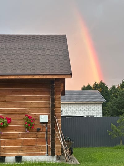The image captures a rustic scene with a prominent wooden structure in the foreground, possibly a house or shed, featuring horizontal wooden logs and a dark, textured shingle roof. A white electrical box is mounted on the exterior wall of this structure. Two potted rose bushes with vibrant red flowers are visible on the left side of the wooden building. In the background, a partially constructed building made of white cinder blocks is seen, with a dark, corrugated metal fence separating it from the foreground. A wheelbarrow and some gardening tools are positioned near the fence. Overcast skies are present, but a distinct rainbow arcs across the sky, its colors appearing muted due to the lighting conditions. Lush green grass covers the ground in the foreground and middle ground. The location is identified as Тумілавіцкі сельскі Савет, Belarus.