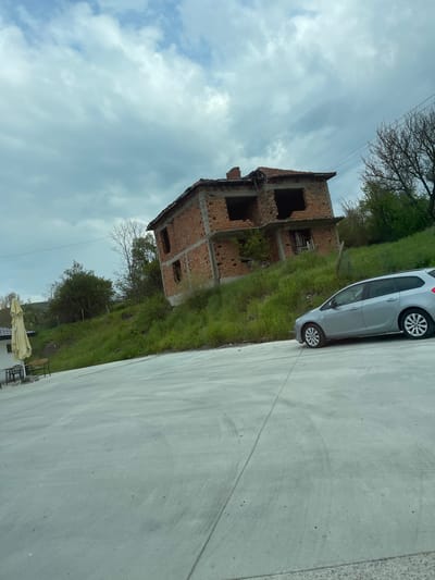 A daytime outdoor scene in Madrets, Bulgaria, captures a static view of a landscape featuring a parked car and a brick structure under a cloudy sky. A light gray compact car is positioned on a flat, gray concrete surface in the middle-right of the frame, facing left. Adjacent to the concrete, a lush green grassy slope ascends, upon which stands a two-story brick building. This structure appears to be either an unfinished construction project or a derelict/damaged house, characterized by exposed red brickwork and empty window and door openings.
The sky above is filled with soft, diffused clouds, suggesting an overcast or partly cloudy day. Bare branches of a tree are visible against the sky in the upper left, while other green trees and foliage dot the grassy hillside. Thin power lines can be seen stretching across the upper part of the image. In the lower left corner, a cream-colored market umbrella and a few dark outdoor chairs are partially visible on another paved area, hinting at a nearby seating arrangement. No individuals are present in the photograph.