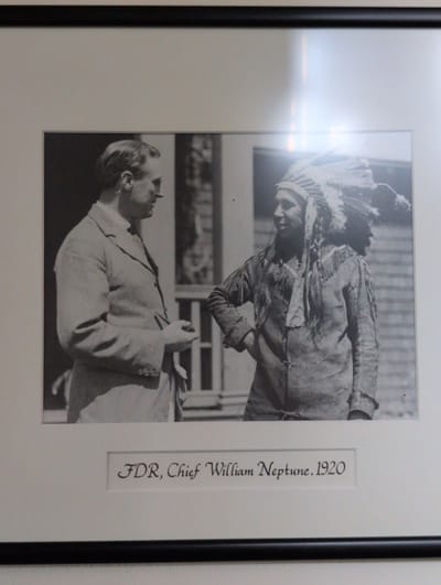 The image is a framed black and white photograph showing two men, one in a suit and the other wearing a Native American headdress and traditional clothing. The man in the suit is on the left, with his side to the camera, wearing glasses. The man on the right is facing the camera, wearing a feathered headdress, a fringed tunic, and has his hand on his hip. The background appears to be a building with windows. Below the photograph, a caption reads "FDR, Chief William Neptune. 1920". The photograph is framed with a dark border. The setting is Campobello Parish, Canada.
