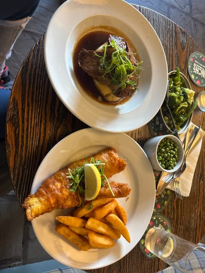 The image depicts a table at a restaurant in West Felton, United Kingdom. Two plates of food are featured. The plate on the left contains a portion of meat, likely braised beef, served on a bed of mashed potatoes and covered in gravy, garnished with sprouts. The plate on the right contains fish and chips, with a lemon wedge and sprouts. Beside the plates are a bowl of peas, a metal dish of broccoli, cutlery on a napkin, and a glass of water with a lemon slice, all arranged on beer mats. The table has a dark, patterned surface. A portion of clothing and a shoe are visible in the lower left corner.