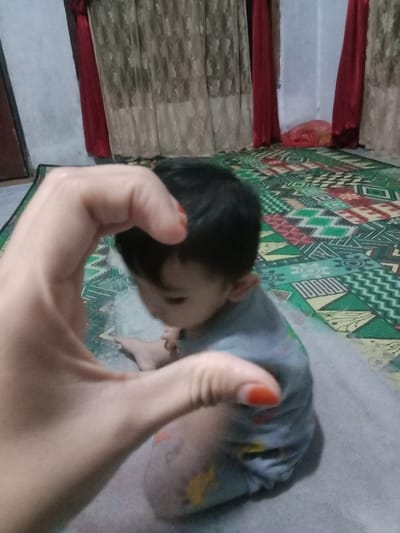 An image captures an indoor scene in the city of Lam Ujong, Indonesia, featuring a child and a prominent foreground hand. In the immediate foreground, a light-skinned hand, with orange nail polish on the thumb and index finger, is held up, forming a partial heart or circular shape. The hand is out of focus. In the midground, a young child, appearing to be an infant or toddler with dark hair, is seated on a light-grey mat or blanket. The child wears a light-colored, long-sleeved garment with multi-colored patterns, including yellow and green. The child's face is turned downwards and to the right, obscuring facial features. The child's bare feet are visible. The floor beneath the grey mat is covered by a patterned mat featuring geometric designs in green, red, brown, and white. In the background, two sets of curtains are visible: a sheer, patterned light-brown curtain overlaid with solid red curtains on the sides. A dark doorframe is present on the left, and a white wall section is visible on the right. A crumpled red plastic bag is situated on the floor in the background, near the right red curtain.