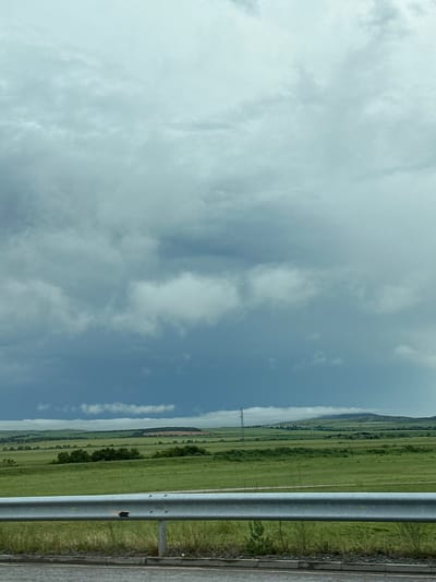 The image presents a broad rural landscape under a heavily clouded sky, viewed from a roadside perspective. Approximately two-thirds of the frame is dominated by the sky, filled with dense, diffuse clouds that vary in shade from light greyish-white to darker blue-grey, suggesting an overcast or possibly pre-stormy day.
Below the expansive sky, a vast stretch of vibrant green fields extends into the distance, characterized by subtle variations in color and texture. A solitary, slender electrical transmission tower or pylon is visible in the middle distance, piercing the horizon line. Further back, low-lying hills or mountains are discernible, appearing somewhat hazy and partially obscured by the atmospheric conditions.
In the immediate foreground, a sturdy, silvery metal guardrail runs horizontally across the right side of the image, indicating the edge of a road. To the right of the guardrail, a narrow strip of asphalt road surface and a grassy verge are visible. The overall scene suggests a journey through open countryside.
The weather is cloudy, and the time of day is daytime, with no direct sunlight casting distinct shadows. There are no people or specific activities depicted. No visible text is present in the image. The setting is consistent with a rural landscape in Vodenichane, Bulgaria.
