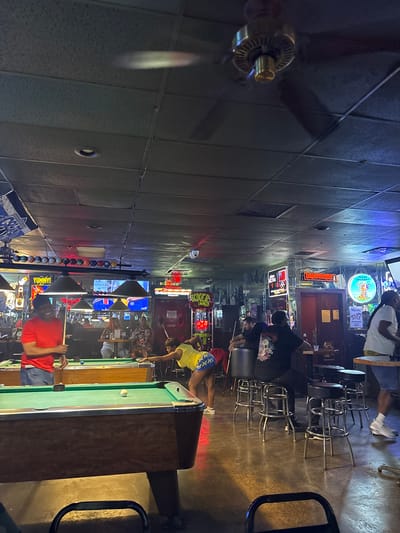 The image shows the interior of a bar in Grand Prairie, United States. People are playing pool on multiple pool tables. Several people are seated at the bar. There are beer signs and sports playing on televisions. The lighting is low and there is a dropped tile ceiling.