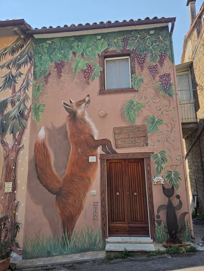 The image depicts a mural on the exterior wall of a building in Viterbo, Italy. The mural features a painted red fox standing on its hind legs, looking upwards towards a window framed by painted grapevines. Below the window, a wooden sign reads "LA VOLPE E L'UVA" (The Fox and the Grapes). To the right of the door, a painted black cat holds a bunch of grapes. The building's facade is painted in warm earth tones, with a wooden double door serving as the entrance. The mural extends to the left, showing a painted tree with branches and foliage. The setting appears to be a residential area, with a narrow street or alleyway in the foreground. The sky is clear and blue.