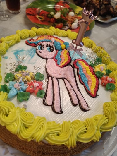 A close-up, overhead shot shows a decorated cake with a pink unicorn figure made of frosting. The unicorn has a rainbow-colored mane and tail and is adorned with small, colorful flowers and silver sprinkles. A rose-gold "1" candle with a crown topper is placed on the cake. The cake is surrounded by yellow frosting piped in a decorative pattern. The background shows a blurred table setting with plates of food and drinks, suggesting a celebration. The image is set in a domestic environment in the city of Pervomayskoye, Russia.