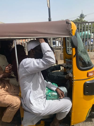 The scene, located in Rijiyar Lemo, Nigeria, depicts the interior and immediate exterior of a yellow commercial tricycle, likely a keke napep, under strong daylight conditions. A man occupies a passenger seat, dressed in a light grey striped long-sleeved tunic and trousers, a white patterned skullcap, and dark sunglasses. He holds the brown canvas canopy frame with his left hand; a green plastic bag containing unspecified items rests on his lap. The tricycle's driver, attired in dark clothing, is partially visible operating the handlebars. Additional passengers are present in the rear compartment, including an individual in a light brown garment and another with a reddish-brown head covering. The background features a metal fence, green foliage, and a prominent light-colored mosque minaret topped with a green dome and a crescent moon finial.