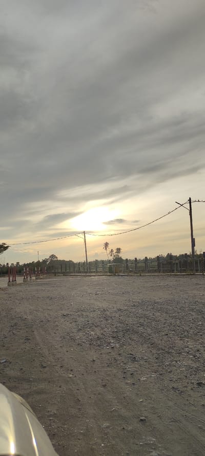 This image depicts an outdoor scene at dusk in Ranto, Indonesia. The sky is filled with dramatic, heavy grey clouds, but a bright, diffused sunlight breaks through them on the horizon, casting a warm, golden glow. This suggests it might be sunrise or sunset. The foreground is a rough, gravelly ground, possibly a dirt road or parking area, with tire tracks visible. In the mid-ground, there is a barrier made of metal posts and rails, with several red and white marker posts lining a path to the left. Beyond the barrier, a few palm trees stand tall against the sky, and dense vegetation forms a natural backdrop. The overall impression is one of a serene, possibly rural, landscape at the end of the day. There are no people visible in the image, but a faint reflection of what appears to be the hood of a car is present in the bottom left corner. The setting appears to be a transitional area, perhaps near a road or a pathway, with construction or maintenance markers suggesting some ongoing activity or designated zone.