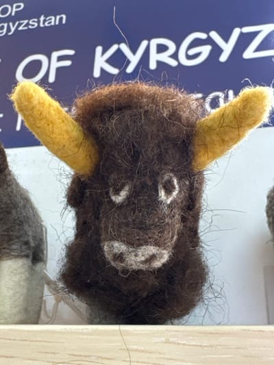 The image displays a close-up of a handcrafted, felted animal head, likely a yak or bull, positioned vertically. The animal's head is primarily dark brown, characterized by a shaggy and coarse texture composed of distinct fibers. It features two prominent, upward-curving yellow horns. Simple white circular markings serve as eyes, and a larger white, textured area forms the snout or muzzle.

The felted head is set against a vivid blue background, which appears to be part of a sign or informational display. White capital letters are clearly visible on this blue surface, spelling "OF KYRGYZ." Further down, partially visible text includes "OP" and "gyzystan," strongly suggesting the complete phrase is "SHOP OF KYRGYZSTAN." To the right of the animal head, a light-colored wooden or laminated surface is visible, possibly part of a display shelf or frame. A section of another felted object, featuring grey and white tones, is partially visible at the bottom of the frame. The image captures the fibrous detail of the felted material, emphasizing its artisanal quality. The visible text and the provided location context of Кашат, Kyrgyzstan, indicate that the item is a local craft or souvenir.