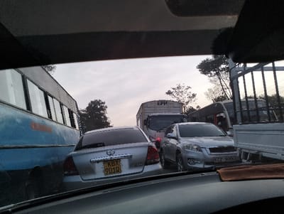 The image provides a view from the interior of a vehicle, looking out through the windshield at a road with heavy traffic. The top and bottom edges of the frame show parts of the car's interior, specifically the roof lining at the top and the dashboard with a brown cloth on the bottom right.

The scene depicts a traffic jam, with multiple vehicles stationary or moving very slowly. The sky appears overcast or hazy, suggesting either an early morning, late afternoon, or evening setting.

From left to right, the visible vehicles include:
*   On the far left, a large, old-looking blue bus, with several rectangular windows along its side. Its front is out of view.
*   In the immediate foreground-middle, the rear of a silver sedan is visible. A Toyota emblem is on its trunk. Its license plate reads "KBX 901Q".
*   Just behind the sedan and slightly to the right, a white box truck with "EPH" printed on its side is visible, facing the viewer.
*   To the right of the sedan and slightly forward, a silver SUV (resembling a Toyota RAV4) is seen from the front. Its license plate reads "KCZ 900U".
*   On the far right, parts of another bus, possibly white or light-colored, are visible, partially obscured by the metal frame of what appears to be a white pickup truck or a flatbed lorry.

Trees and foliage are visible in the background, interspersed between and behind the vehicles, suggesting a roadside lined with vegetation. The overall impression is one of congestion and waiting. The license plate formats "KBX 901Q" and "KCZ 900U" are characteristic of Kenyan registration plates.