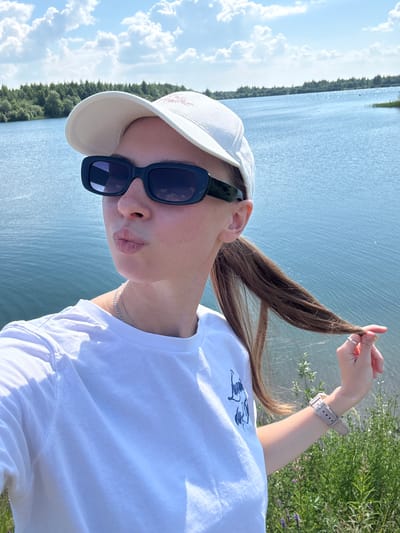 The image depicts a young woman taking a selfie outdoors with a lake and trees in the background. She is wearing a white baseball cap, dark rectangular sunglasses, and a white t-shirt with a graphic print. Her lips are pursed, and she is holding a strand of her long brown hair in her right hand. A silver necklace is visible around her neck, and she is wearing a light-colored watch on her left wrist. The environment is natural, with a body of blue water, a line of green trees along the horizon, and a bright blue sky with white clouds. Green vegetation and some small purple flowers are in the foreground. The provided context suggests the location is the city of Zaosinovo, Russia.