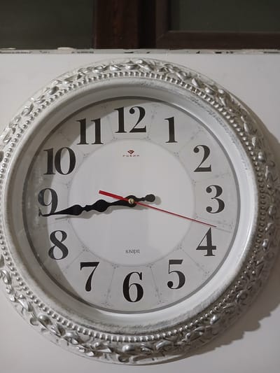 This is a close-up, eye-level photograph of a white, ornate wall clock with a silver-toned, decorative frame. The clock face is white with large, black Roman numerals and ornate grey markings. The clock has three hands: a black hour hand, a black minute hand, and a thin red second hand. The hour hand is pointing slightly past the number 5, the minute hand is pointing slightly past the number 6, and the second hand is pointing at the number 4. This indicates the time is approximately 5:32. On the clock face, in smaller text, the word "РУБИН" is visible near the 12, and "кварц" is visible near the 3. The clock is mounted on a plain white wall, with a dark framed window or doorway visible on the left edge of the frame. The lighting suggests an indoor setting, possibly in the evening or with artificial light, as evidenced by the shadows cast by the clock's decorative frame. The overall impression is of a decorative, functional timepiece in a domestic or public space. The location context is Ijevan, Armenia, though this is not explicitly visible in the image itself.