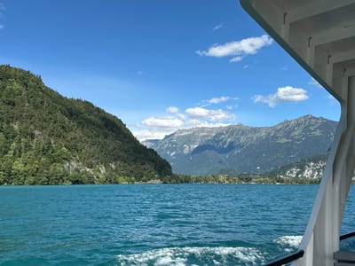 The image captures a scenic view of Lake Thun near Sundlauenen, Switzerland, from the perspective of a moving boat. The turquoise water of the lake fills the foreground, with a wake trailing behind the unseen vessel. On the left, a densely forested mountain slopes down to the water's edge. Across the lake, a distant shore is lined with trees and buildings, and beyond that, a range of majestic mountains with rocky peaks dominates the background under a clear blue sky dotted with white clouds. The right side of the image shows a portion of the boat's white structure, including a railing and part of its upper deck, framing the expansive landscape.