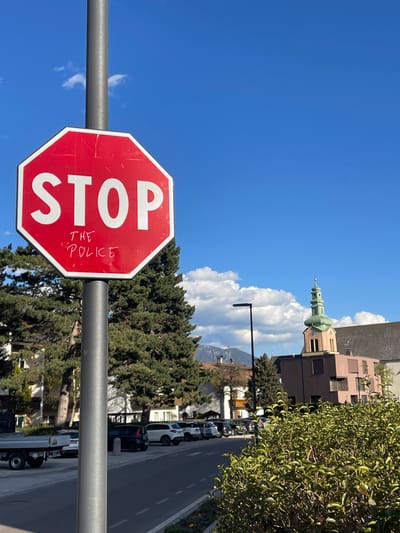 A low-angle shot of a stop sign defaced with the words "THE POLICE," attached to a horizontal silver pole. In the background, an Italian cityscape, with a building with a tower, puffy white clouds, and a bright blue sky. A road with parked cars is partially visible. The photo likely captured in Sterzing - Vipiteno, Italy, judging from the architecture.
