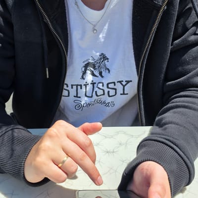 A person's torso and hands are visible, seated at a table with a patterned surface. They are wearing a black zip-up hoodie over a white t-shirt with a black graphic print that includes a lion and the words "STUSSY Sportswear". A silver necklace with a heart-shaped pendant is worn around their neck. The person is holding a smartphone and appears to be interacting with it, with one hand resting on the table and the other finger pointing downwards towards the device. A gold ring is visible on the ring finger of the left hand. The setting appears to be outdoors, with bright sunlight casting shadows. The exact location in Cannon Beach, USA is not specified.