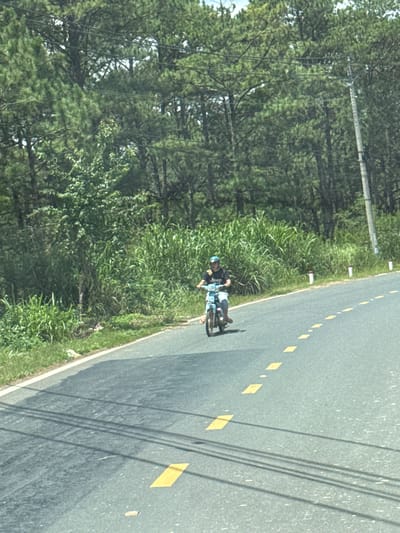 The image depicts a person riding a light blue motorcycle on a paved road. The rider is wearing a dark t-shirt with white lettering that appears to spell "Jeep" and a light-colored helmet. The motorcycle has a license plate visible. The road is marked with yellow dashed lines. To the left of the road, there is a grassy embankment with tall green vegetation, and beyond that, a dense forest of pine trees. To the right of the road, there are several white bollards with red tops, and a utility pole with wires. The scene appears to be in a rural or suburban area, possibly Thôn 6, Vietnam, given the vegetation and road markings. The lighting suggests it is daytime.