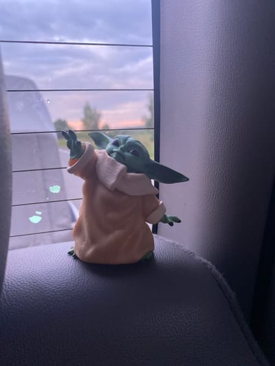 An action figure of Grogu, also known as Baby Yoda, is positioned on the textured dark gray headrest of a car seat. The figure is depicted in a light tan robe with a cream-colored collar. Its left arm is extended upwards and to the left, with its index finger pointing. The figure's head is turned to its right, with large ears and green skin.
Behind Grogu, a car window is visible, featuring horizontal defroster lines. Through the window, a soft, cloudy sky is visible, with the sky transitioning to soft pink and orange hues near the horizon, suggesting either sunrise or sunset. Sparse trees can be seen in the distance. There are two smudges of light green on the window, near the bottom left of the figure. The right side of the image is dominated by the textured dark gray upholstery of the car's interior. There is no discernible information about the location of Balakhi, Russia within the image.