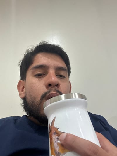 An adult male with dark hair and a short beard is depicted in a close-up, chest-up shot, presumably indoors in Adrogué, Argentina. He is wearing a dark blue garment. His gaze is directed towards the camera. The subject is holding a white insulated tumbler with a silver-colored rim in his right hand, positioned in front of his face. His lips are pursed and in contact with the rim of the tumbler, indicating an act of drinking. An illustration of a reddish-brown animal's head, resembling a fox, is visible on the side of the tumbler. The background consists of a plain, light-colored wall.
