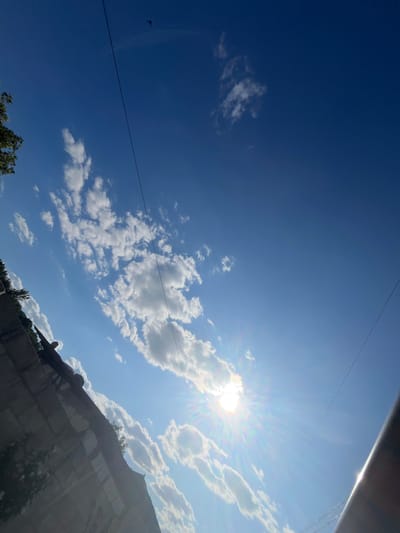 The image captures a bright, sunlit sky with scattered cumulus clouds. In the foreground, a weathered concrete structure runs diagonally across the lower left portion of the frame, with some green foliage growing on its surface. A power line stretches across the sky from the upper left towards the center. The sun is prominently visible in the lower right quadrant, emitting strong rays and lens flare. A person is partially visible standing on top of the concrete structure. The scene appears to be an outdoor urban environment, possibly in Telenești, Moldova, given the context.
