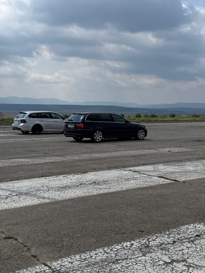 The image shows two cars, a white station wagon and a dark-colored station wagon, parked on a cracked asphalt surface with faded white lines, likely a road or parking area. In the background, there is a view of distant hills or mountains under a cloudy sky. The location is Baiachevo, Bulgaria, based on the license plate "B" prefix, which is specific to the region of Burgas, Bulgaria. The scene appears to be in a rural or semi-rural area.