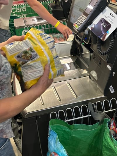 The image depicts a transaction at a self-checkout station within a supermarket located in Esperance, Australia. In the foreground, a person with light skin, wearing a short-sleeved patterned shirt and blue jeans, is holding a large yellow and white flexible package, likely a frozen food item with a circular image of a prepared dish, over the reflective metal scanning surface. Their hands are positioned on the package as if in the process of scanning or placing it. To the right and further back, a second person, wearing a light-colored top and a yellow wristband on their right wrist, extends their arm towards a white plastic bottle and other indistinct grocery items on the adjacent staging area.

The self-checkout machine features a black frame, a vertical digital display screen, and a payment terminal equipped with a keypad and card reader. Attached to the payment terminal is a promotional display advertising "Apple Pay," featuring an image of an iPhone screen displaying "9:41." A black coiled cable extends from the side of the payment terminal. Below the scanning surface, a chrome-plated metal rack supports a bright green reusable shopping bag, with a segment of another multi-colored bag visible to its right. The background shows a green vertical surface marked with "Card Only," indicating another checkout lane or a partition. The scene is uniformly illuminated by overhead lighting.