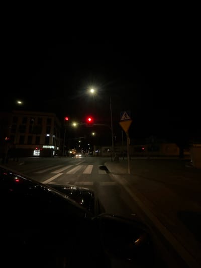 This is a night-time, street-level, wide-angle shot from inside a vehicle looking towards a city intersection. The foreground is dominated by the dark silhouette of the car's hood and windshield. The middle ground features a crosswalk with white stripes on a dark asphalt road. Across the intersection, a multi-story building with illuminated windows is visible on the left, and a sign that reads "JUBLIER" is also present. Traffic lights are illuminated with red signals. Streetlights cast a glow on the scene. A pedestrian crossing sign is visible on the right side of the image. The background is dark, with some distant lights and the silhouette of trees. The scene is likely in Siedlce, Poland, given the context provided.