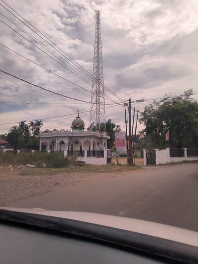 The image presents a daytime street view in Paya Tringgadeng, Indonesia, captured from inside a vehicle. A white mosque with a green and orange dome, featuring an Islamic crescent finial, stands prominently on the left side, surrounded by a black metal fence. Palm trees are visible behind the mosque.

To the right of the mosque, a tall, metallic lattice communication tower stretches high into a cloudy, overcast sky. Numerous power lines traverse the upper half of the image. A wooden utility pole is situated near the center-right, bearing a "Fresh Laundry" sign. This sign displays "Fresh Laundry" in English and detailed information in Indonesian, including "Mulai Rp. 6000" (Starting Rp. 6000), "Diterima / Diambil" (Received / Taken), "Buka Tiap Hari" (Open Every Day) with hours "08:00 - 22:00", a phone number "0852...", and the location "Jl. Panglima Polem, Paya Jaya".

Further right, large green trees and two red and white Indonesian flags are seen near a white wall and gate. The foreground shows a paved road, with the bottom portion of the image revealing the dashboard and windshield of the car from which the photograph was taken. The ground adjacent to the road and the mosque's fence is somewhat unkempt with dry vegetation and scattered litter. No people are visible in the scene.