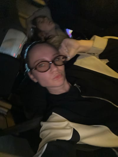 The image depicts two individuals within a dimly lit interior, located in Çiftlikköy, Turkey. In the immediate foreground, a female subject, with fair skin and dark hair pulled back from her face, directly observes the camera. She wears dark, oval-framed eyeglasses, a small hoop earring in her left ear, and a nostril piercing. Her lips are slightly pursed. She is dressed in a black top or jacket featuring a distinct cream or beige panel extending across the right shoulder and arm. A ring is visible on her right hand, which is positioned near her right temple. In the upper background, partially obscured behind the foreground subject, a second individual is visible. This person, consistent with female characteristics and lighter hair, is seen with their head tilted backward, indicating a state of repose. Their attire appears to be light-colored. The surrounding environment is characterized by profound darkness, with minimal discernible features. Vague, multi-colored light sources are evident in the upper-right quadrant, suggesting artificial illumination or reflections within a confined space.