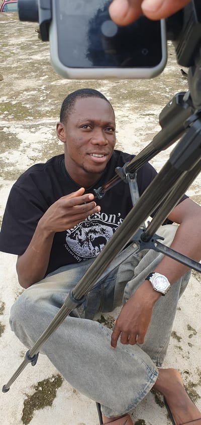 A young Black man is sitting on a concrete surface, facing slightly to his right and looking towards the camera with a gentle smile. He is wearing a black t-shirt with a graphic print featuring animals and text, and light-wash denim jeans. His left hand is raised, with fingers slightly spread, as if in the middle of speaking or gesturing. A tripod is positioned in front of him, with some of its legs visible. He is wearing a watch on his left wrist. The ground appears to be a light-colored, rough concrete surface, possibly outdoors. In the background, a portion of a red vehicle is visible. The overall scene suggests a casual, possibly creative setting, given the presence of the tripod, suggesting filming or photography. The location is stated to be in Anguldi, Nigeria.