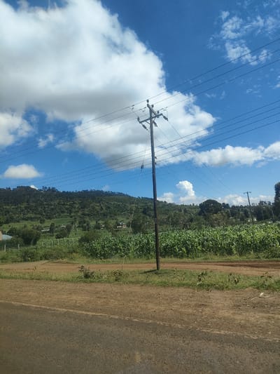 The image depicts a rural scene in Kibirichia ward, Kenya. A wooden utility pole with multiple power lines is prominently positioned in the foreground. Behind the pole, a field of maize or a similar crop stretches across the landscape. In the distance, a hill covered in trees is visible under a partly cloudy blue sky. The foreground shows a dirt road with some vegetation along its edges. There are no people visible in the image.