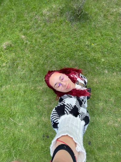 A medium-angle, full shot shows a woman lying on a patch of green grass. The location is unknown; there is no evidence to support it being in Barkach, Bulgaria. The woman has red hair and is wearing glasses. She is wearing a black and white patterned sweater. Her right arm is extended towards the camera, with a black and red strap visible on her wrist. The background is entirely green grass.