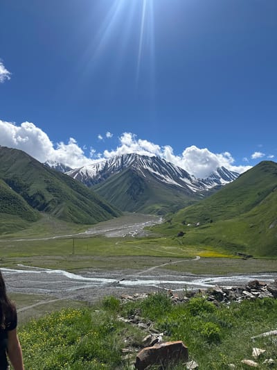 The image shows a vast, mountainous landscape in Georgia under a bright blue sky with sunbeams and scattered clouds. The foreground features a person's shoulder and back on the left, with green grass and yellow wildflowers. A rocky riverbed or dry river snakes through the valley, with green, rolling hills and mountains on either side. The distant peaks are capped with snow, creating a stark contrast with the lush green slopes. A small, reddish-brown building is visible in the mid-ground on the right side of the valley, with a winding dirt road leading towards it. Power lines and poles are present in the valley floor, suggesting some human presence and infrastructure. The overall scene depicts a serene and majestic natural environment.