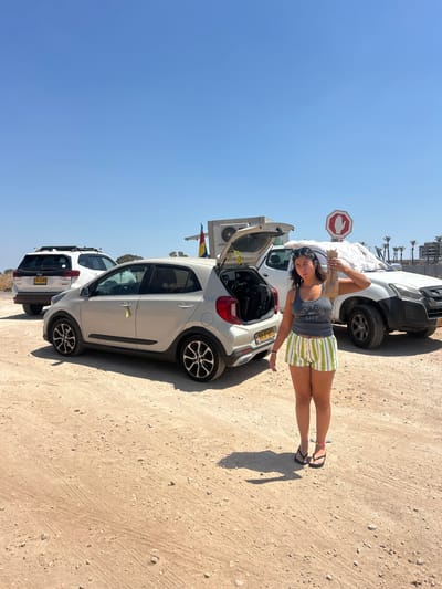 The image displays an outdoor scene under a clear, bright blue sky, located in the Galil Tachton Regional Council, Israel. The ground consists of an unpaved, light brown dirt and gravel surface with visible tire tracks.
One female individual stands in the mid-foreground, positioned slightly right of the camera's center. She possesses dark hair, with sunglasses resting on her head. She wears a grey tank top with "LIVE YOUR LIFE" partially visible, green and white horizontally striped shorts, and black flip-flops. Her right arm is bent, holding an elongated, light-colored object, appearing cylindrical and wrapped in material, held vertically near her head. Her left arm hangs beside her body.
Three vehicles are parked on the dirt surface. To the left of the individual, a light grey Kia Picanto hatchback (Israeli license plate 403-12-801) is parked facing right. Its rear hatch is open, revealing dark-colored objects inside. A yellow ribbon is tied to its driver's side door handle.
To the right of the Kia Picanto, a white pickup truck (Israeli license plate 313-57-601) is parked, also facing right. A light-colored, wrinkled material covers part of its front hood. Behind this truck, a red octagonal stop sign featuring a white hand symbol is visible. Adjacent to the stop sign, an external air conditioning unit is mounted on a structure, and a flag with horizontal stripes of yellow, red, and green hangs from a nearby pole.
In the background, to the left of the Kia, a white Subaru Forester SUV (Israeli license plate 729-79-601) is parked, facing right, equipped with a black roof rack.
The distant background includes a line of palm trees and multi-story buildings visible on the horizon to the right, indicating some form of development beyond the immediate unpaved area. Sparse, dry vegetation is present in the middle ground on the left.