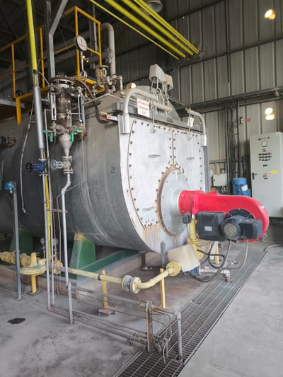 The image shows a large industrial boiler, likely a steam boiler, within a factory or power plant setting. The boiler is primarily silver-gray and cylindrical, with various pipes, valves, and gauges attached. A prominent red and black burner unit is affixed to the front of the boiler. Yellow pipes and supports are visible throughout the scene, along with a metal control panel with buttons and indicators. The floor is concrete, with a metal grate area in the foreground. The overall atmosphere suggests an industrial environment.
There are no people visible in the image. The activity appears to be stationary, with the boiler likely in a standby or operational state. No specific time of day or weather conditions are discernible. The setting appears to be an indoor industrial facility with metal walls.
Visible text includes Hebrew characters on a white sign affixed to the boiler, which appears to read "דוד חימום" (heating boiler) and a number "# 2". Another sign with similar text is partially visible. On the control panel, there are also labels and indicators, some of which may be in Hebrew or English, but they are not legible enough to read. The number "2" is also painted on the side of the boiler. The location context provided is Dimona, Israel, which aligns with the presence of Hebrew text.