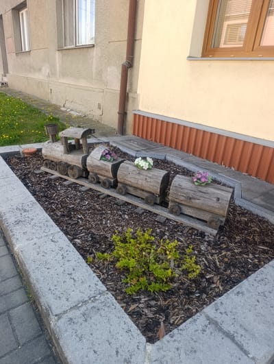 In the city of Výkleky, Czechia, a charming garden feature depicts a wooden train set placed in a flowerbed. The train, crafted from logs, consists of a locomotive and multiple cars, each adorned with colorful flowers. The flowerbed is filled with dark mulch and bordered by concrete slabs. To the left, a paved walkway and a grassy area with dandelions are visible, while a building with windows and a downspout stands behind the garden.
