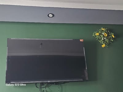 This image captures an interior scene within a structure in Dobroslavtsi, Bulgaria. The room features a dark green wall, surmounted by a horizontal grey band, which transitions into a white ceiling. A single, circular recessed spotlight, black-rimmed, is centered within the grey ceiling band. A large, black flat-screen television is mounted on the dark green wall, positioned left of the frame's center. Clear protective film or tape is visible along the top, bottom, and side edges of the television screen. A small, square red-orange sticker is affixed to the upper right corner of the television's frame. The brand "TCL" is faintly legible on the bottom right bezel. A bundle of black cables hangs beneath the television against the wall. To the right of the television, a decorative floral arrangement consisting of green foliage and multiple yellow flowers is mounted on the dark green wall. The image includes a "Galaxy S23 Ultra" watermark in the lower-left corner. No individuals are present.