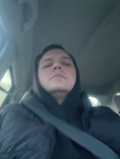 A person with fair skin is in a car, wearing a dark hooded sweatshirt and a seatbelt. The interior of the car is visible, with a gray roof and a glimpse of the outside seen through the windows. The location is not discernible as Maksimkovo, Russia based on the provided image.