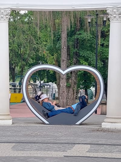 An unidentified person, wearing a white hat and blue denim jacket over a pink shirt, reclines on a grey, heart-shaped bench. The person is holding a mobile phone and appears to be looking at it. The bench is situated outdoors, framed by two white columns and an archway, with a park-like setting in the background featuring trees and foliage. In the distance, there are some playground-like structures and a lamppost. The ground in front of the bench is paved with grey interlocking stones. The scene appears to be a public space, possibly a park or recreational area, in Alikonovka, Russia.