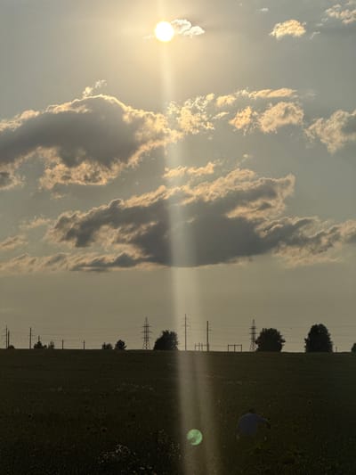 The photograph captures a wide-angle view of a sun-drenched sky over a rural landscape, possibly Aksaitovo, Russia. The sun, a brilliant orb with visible rays, shines through scattered clouds, casting a beam of light downwards. In the middle ground, a silhouette of power lines and transmission towers stretches across the horizon, interspersed with trees. The foreground is a dark, low-lying field, likely agricultural, with hints of vegetation and a single human figure in the lower right quadrant, bent over as if working. The overall atmosphere is one of late afternoon or early evening, with a warm, soft light dominating the scene.