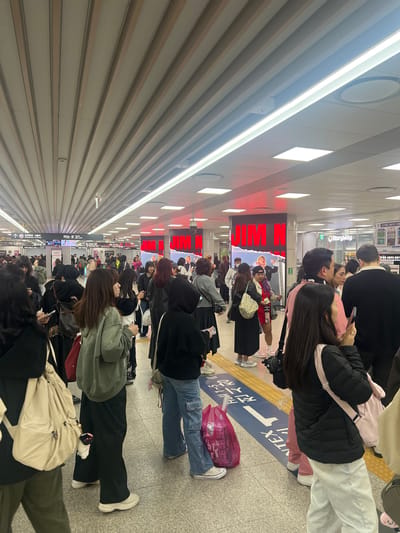 The image shows a crowded scene at a train station in Goyang-si, South Korea. Many people are standing or moving about, seemingly waiting for or exiting a train. Some individuals are using their phones. Signage indicates the direction to Seoul Station, informs about prohibited items such as bicycles, and references transportation cards and chargers. A "JIMIN N" advertisement is visible. The station features a tiled floor, a slatted ceiling with lighting, and tactile paving for the visually impaired.