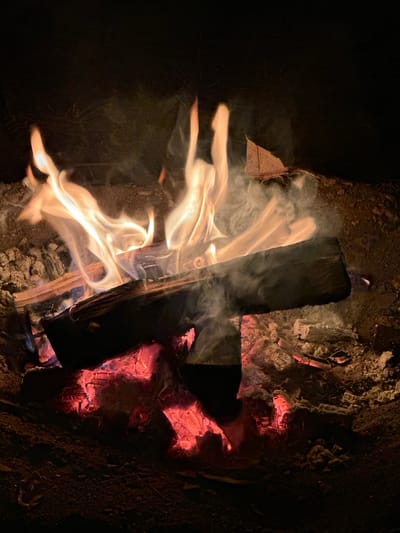 The image depicts a close-up view of a campfire burning in what appears to be an outdoor setting at night. The fire is composed of several logs and glowing embers, emitting flames and smoke. The surrounding area consists of dirt and ash. There are no people visible in the image. The scene suggests a recreational activity such as camping or gathering around a fire. Given the location constraint provided, the scene would be situated in or around the city of Жемчужный, Russia, likely in a park, backyard, or a designated campsite. The lighting is low, with the primary illumination coming from the fire itself.