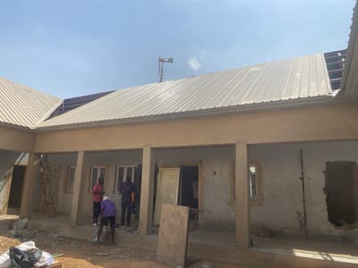 The image depicts a construction site in Bisichi, Nigeria, under a clear blue sky. A single-story, L-shaped building is under construction, with a newly installed corrugated metal roof covering most sections, while one central roof section exposes wooden roof trusses. The building's exterior features a light beige plaster finish on its upper walls and pillars, with the lower walls exhibiting a rough, unplastered grey concrete surface.
Four adult males are visible in the foreground. One male, wearing a purple shirt and dark shorts, is bending down, actively sweeping or clearing debris from the dirt ground in front of the building. To his right, two other males stand near open doorways: one in a red shirt and dark trousers, and another in a plaid shirt and dark trousers. A fourth male is partially obscured behind the individual in the red shirt.
The ground around the building is unpaved, consisting of loose dirt and scattered construction debris, including a black plastic bag and wooden planks. Several window openings are present, some with white frames installed, others bare. Doorways are visible, with one having a light-colored wooden door partially open. A wooden ladder leans against the wall on the far left. In the distant background, above the roofline, a structure with what appears to be a weather vane or antenna is visible against the sky.