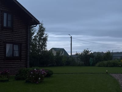 The image depicts a yard in Shebarshino, Russia, under an overcast sky. On the left, a portion of a dark wooden log house is visible, featuring two dark-framed windows. In front of the house, a lush green lawn is dotted with flower beds containing pink and white blooms. Beyond the lawn, various trees and shrubs form a backdrop, with a light-colored house featuring a green roof partially visible amongst the foliage. A utility pole with wires and a street lamp stands further back. The overall scene suggests a rural or suburban setting on a cloudy day.