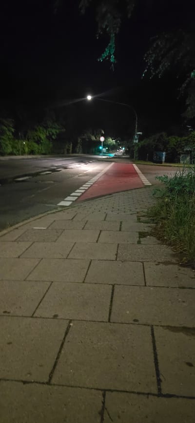 The image captures a nighttime view of a street in Seevetal, Germany, likely near the intersection of Maschener Bahnhofstraße and Glüsinger Straße, based on visual cues. A well-lit crosswalk, painted red with white dashed lines, crosses the asphalt road, which exhibits signs of wear and some potholes. A pedestrian sidewalk, paved with light-colored, rectangular concrete slabs, runs alongside the road on the right. Overgrown grass and vegetation border the sidewalk. In the background, trees and foliage line the street. Traffic signals and streetlights are visible, illuminating the scene. A speed limit sign (30 km/h) is posted on a pole. The overall impression is one of a quiet, ordinary street scene at night, typical of a suburban or semi-urban environment.