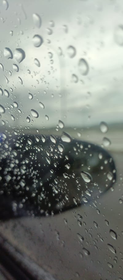 The image presents a close-up, vertical shot of a window covered in numerous water droplets, likely from rain. The droplets vary in size and shape, clinging to the glass, with many exhibiting distinct highlights and reflections that give them a crystalline appearance. They are sharply in focus, creating a textured foreground.
Through the translucent, water-covered glass, the background appears heavily blurred and out of focus. A dark, indistinct shape, possibly a car's side mirror or part of the vehicle's body, is visible towards the lower middle and right side of the frame. Above and beyond this, a soft, grey, and white expanse suggests an overcast sky or a distant, muted landscape.
The scene depicts a rainy or post-rain moment, viewed from inside a vehicle. The lighting is diffused and soft, characteristic of an overcast day. The overall impression is one of quiet contemplation, observing the outside world through a veil of rain. The location context provided is Glee Gogo, Indonesia. There is no visible text in the image.
