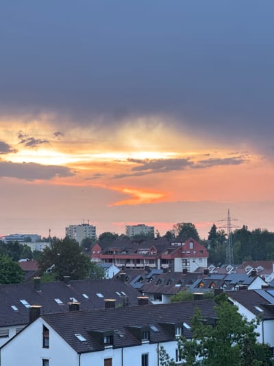 The image shows a panoramic view of a residential area in Sindelfingen, Germany, during a sunset. The sky is filled with dramatic clouds, transitioning from a deep blue at the top to vibrant oranges and yellows closer to the horizon. In the foreground, the dark-tiled roofs of numerous houses with white walls and visible chimneys and dormer windows dominate the scene. Many of these roofs are equipped with solar panels, suggesting a focus on renewable energy. In the mid-ground, a dense line of green trees separates the residential area from taller buildings, including apartment complexes and what appears to be an industrial or commercial structure. A prominent electricity pylon stands tall among the trees on the right side of the image. The overall impression is a suburban landscape bathed in the warm, colorful light of a setting sun.
