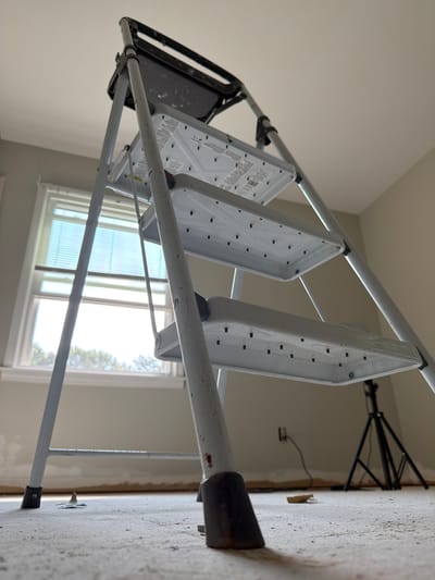 A low-angle, ground-level, medium shot shows a three-step metal stepladder placed in the center of an empty room. The ladder is light gray with a black top platform and black non-slip feet. The floor is covered in a fine white dust, suggesting construction or renovation work. A window with white blinds is visible to the left of the ladder, letting in natural light. In the background, a black tripod with a light fixture is set up on the dusty floor. The walls are painted a neutral, light beige color. The scene appears to be an interior residential space in Stallings, United States of America.