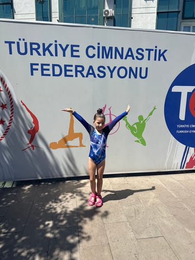 A young girl in a blue and white gymnastics leotard with butterfly patterns and pink sandals poses in front of a large mural on a sunny day. The mural features large blue text that reads "TÜRKİYE CİMNASTİK FEDERASYONU" and stylized silhouettes of gymnasts in red, orange, and green. To the right of the girl, a blue circle contains a large white "T" and smaller text that reads "TÜRKİYE CİM" and "TURKISH GYM". The background is a light-colored building with several windows. The ground in front of the mural is paved with square stone tiles, and the scene is partially covered by shadows from nearby trees. The girl is standing with her arms outstretched, mirroring the pose of one of the silhouettes. The location is identified as Sancaktepe, Turkey.