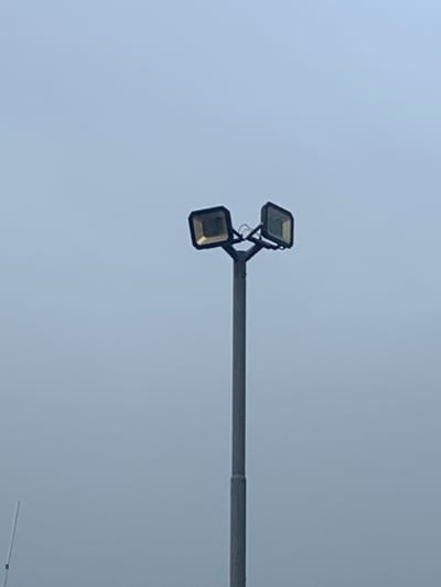 A tall, grey pole supports two square floodlights, one on each side, angled outwards. The floodlights appear to be made of dark metal with illuminated glass fronts. The sky behind is a pale, overcast blue. A thin white pole or antenna is visible in the lower left corner of the frame. The scene is devoid of people or any other significant objects.
