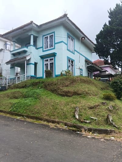 The image displays an exterior view of a two-story residential building, painted light blue with contrasting darker blue trim around windows and along horizontal bands, situated on an elevated grassy mound adjacent to a paved road in Pô Kô, Vietnam. The house features a prominent tiled roof with wide eaves, multiple white-framed windows equipped with grilles, a ground-floor covered porch with white railings and columns, and an upper-level balcony with similar railings.

A utility pole with a circular light fixture extends vertically near the right side of the main house. Further right and slightly behind, a structure with a reddish-brown tiled roof and a dark gazebo is partially visible, alongside a tall, dark green coniferous tree. The grassy mound supporting the house presents a mix of vibrant green and drier, patchy brown vegetation. Several weathered wooden stumps are scattered on the mound near the road.

In the foreground, a dark asphalt road with minor surface cracks runs horizontally, separated from the grassy incline by a concrete curb. An overcast, light gray sky dominates the upper portion of the frame. To the far left, a white cylindrical water tank is visible on the roof of an adjacent white building. No individuals or specific actions are depicted within the scene. The environment appears static and residential.
